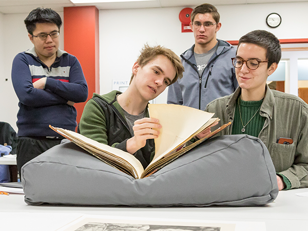 A group of four students gather around an large, aged book that sits atop a big gray pillow. One of the students is carefully turning a yellowed page and peering at its contents.